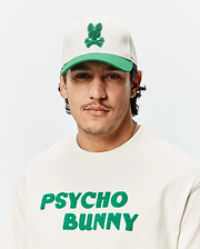 Mens Craig Baseball Cap in off-white with a green brim and embroidered green bunny skull logo, paired with an off-white crewneck sweatshirt featuring a large green chenille Psycho Bunny text logo.