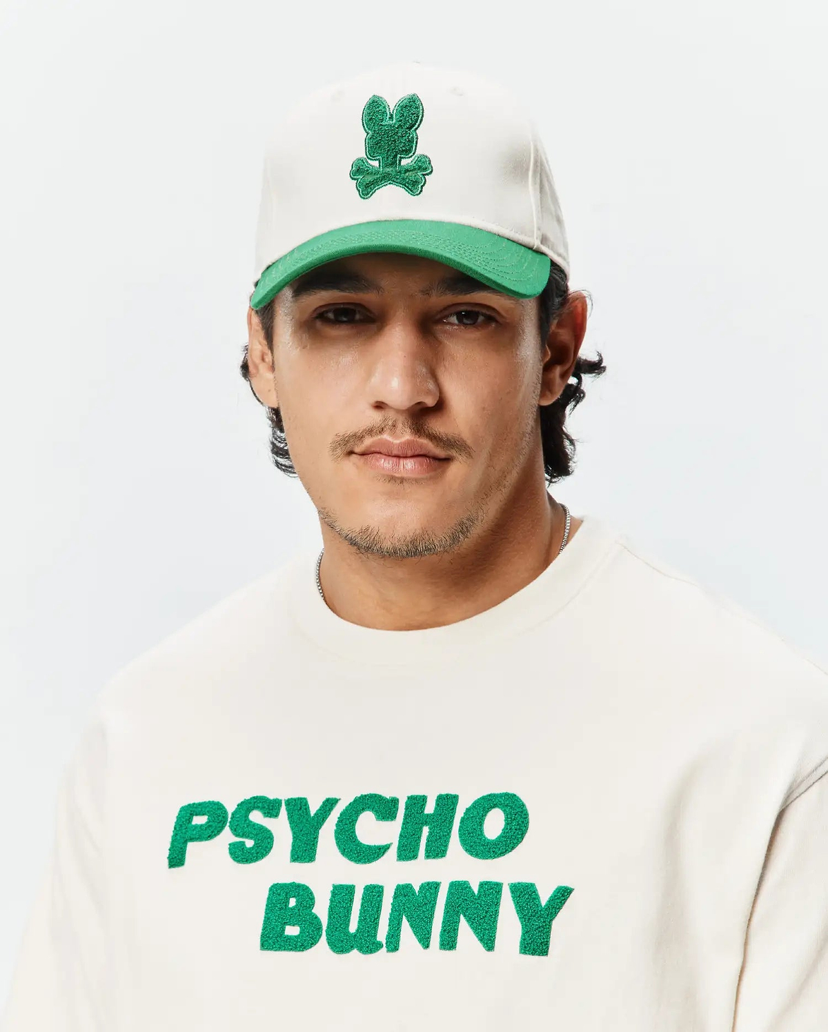 Mens Craig Baseball Cap in off-white with a green brim and embroidered green bunny skull logo, paired with an off-white crewneck sweatshirt featuring a large green chenille Psycho Bunny text logo.
