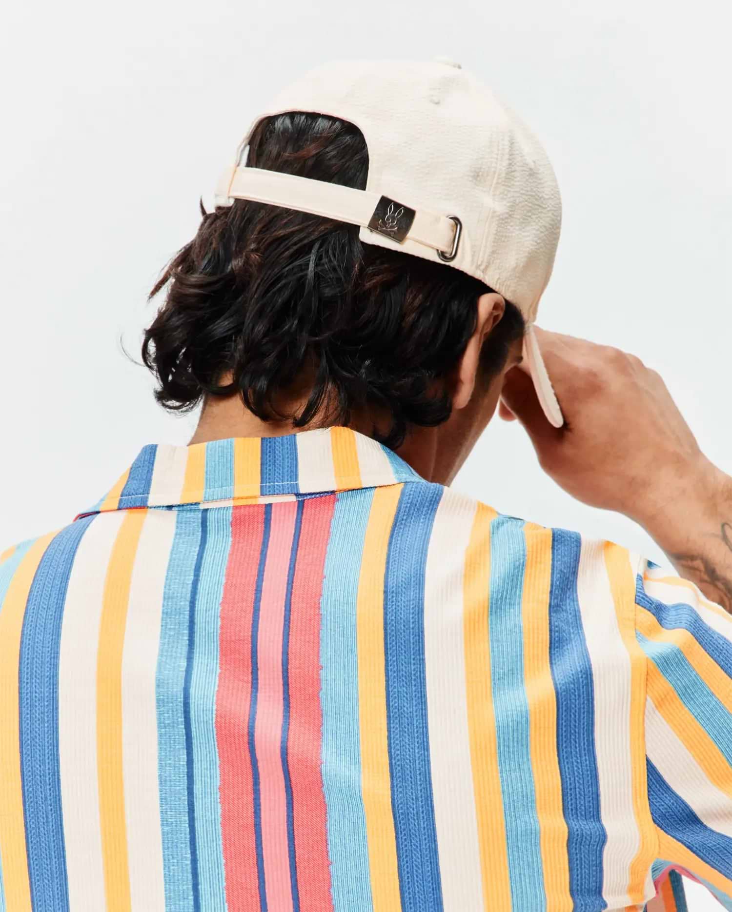 Mens Hubert Baseball Cap in cream textured fabric with a small Psycho Bunny skull logo on the back buckle, paired with a relaxed fit short-sleeve shirt featuring bold vertical stripes in cream, blue, orange, and pink.