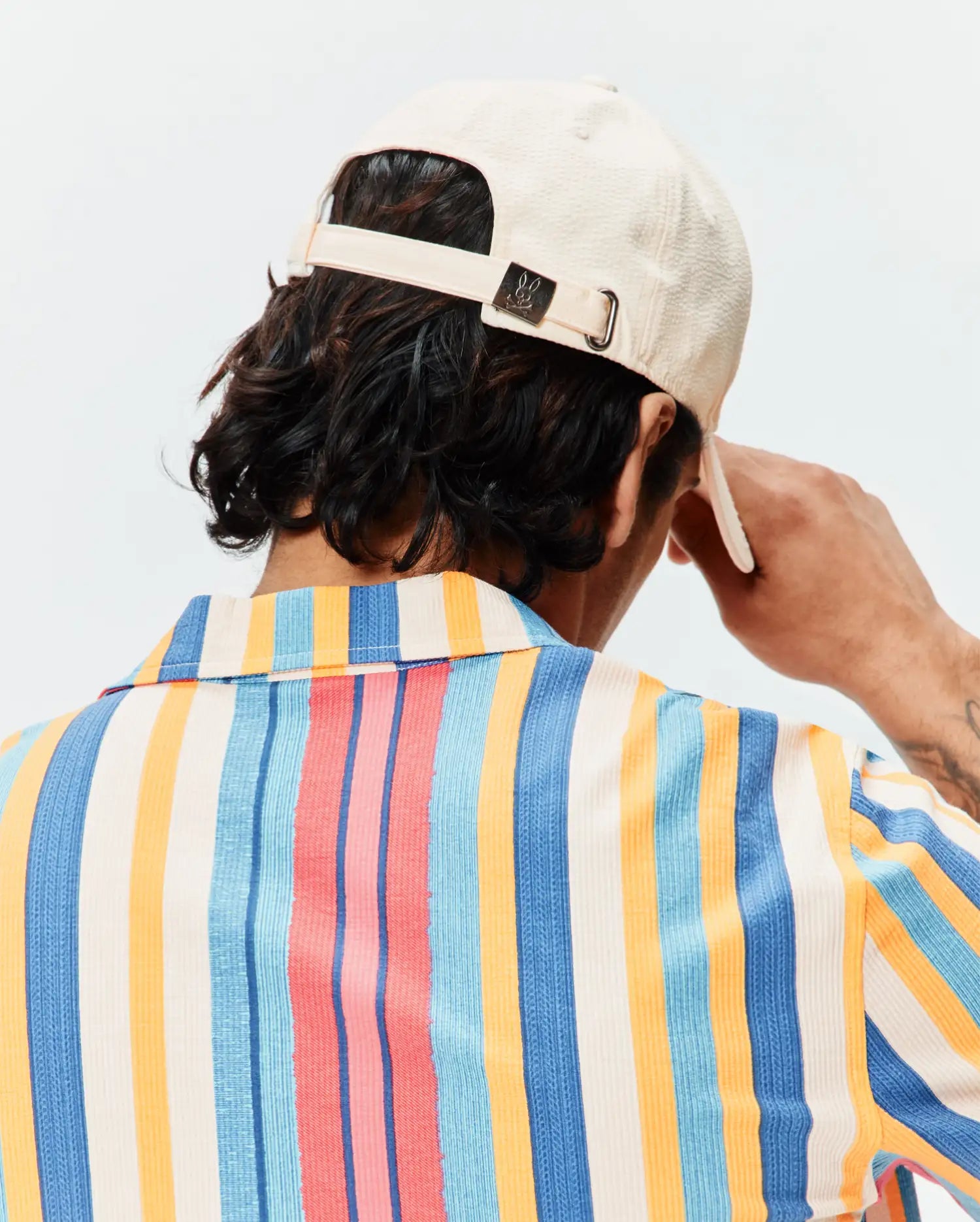 Mens Hubert Baseball Cap in cream textured fabric with a small Psycho Bunny skull logo on the back buckle, paired with a relaxed fit short-sleeve shirt featuring bold vertical stripes in cream, blue, orange, and pink.