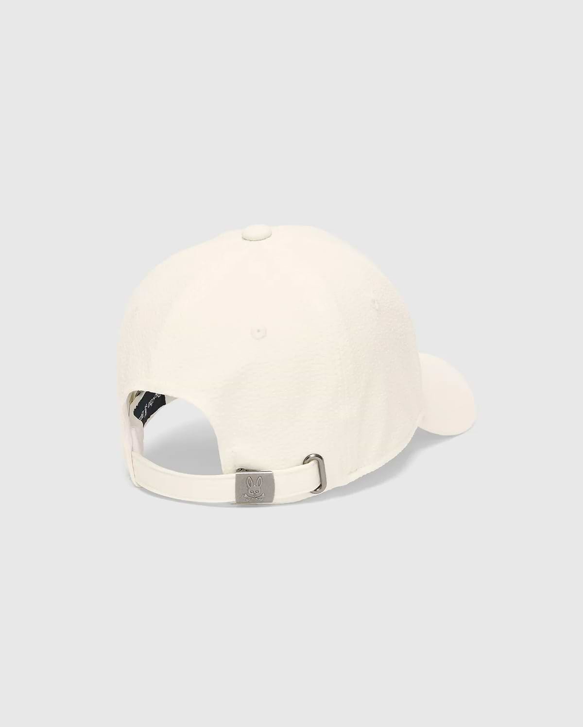 Mens Hubert Baseball Cap in cream textured fabric, featuring an adjustable back strap with a gunmetal buckle embossed with the small Psycho Bunny skull logo and a dark inner band.