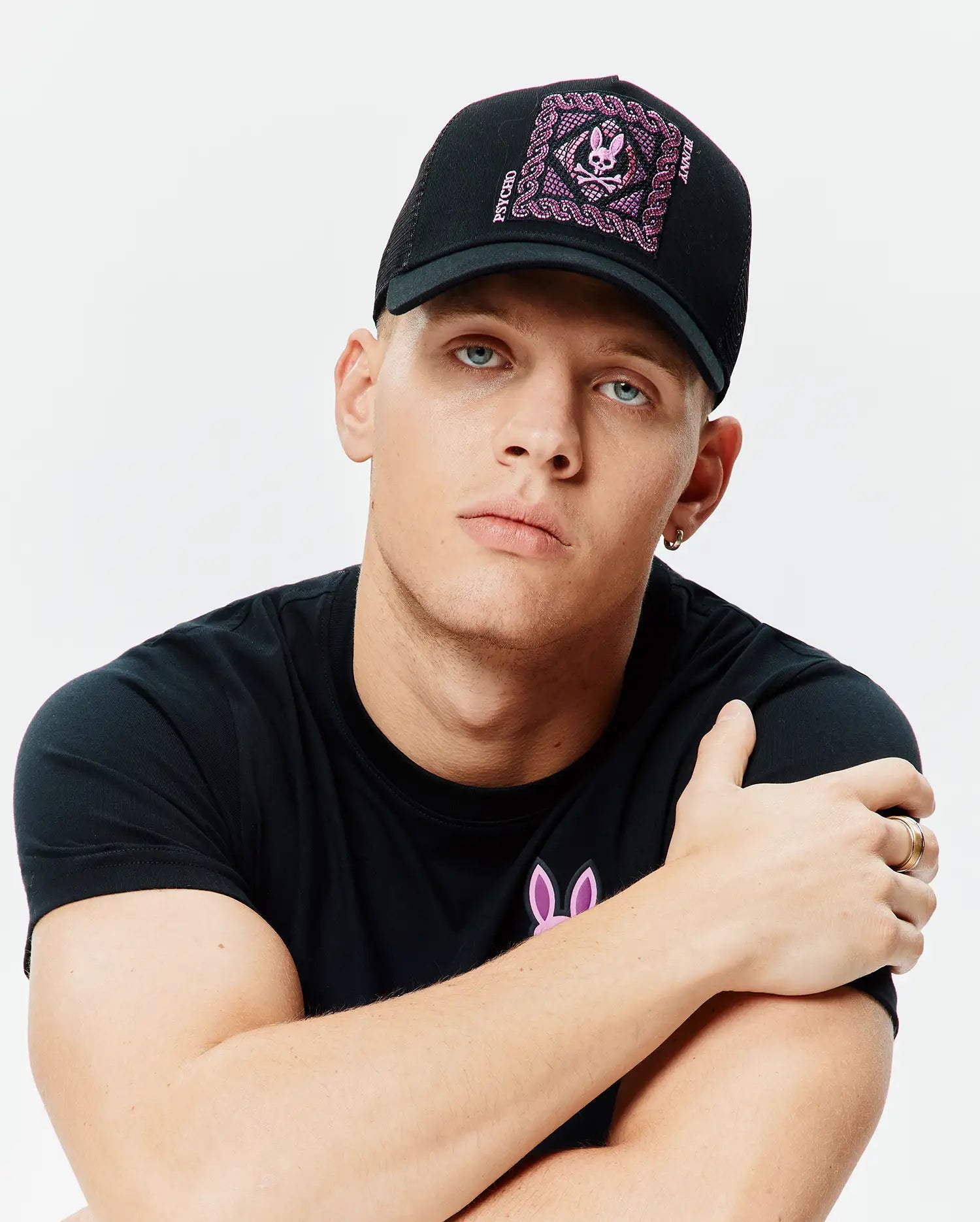 Black Mens Frank Trucker Cap with large pink embroidered Psycho Bunny skull logo and chain detail, paired with a black jersey t-shirt featuring a small pink Psycho Bunny logo on the left chest.