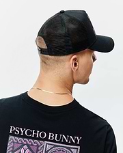 Black classic fit jersey t-shirt with large pink PSYCHO BUNNY text and mosaic graphic on the upper back, paired with a black mesh-back Mens Frank Trucker Cap featuring an embroidered pink Psycho Bunny logo on the front.