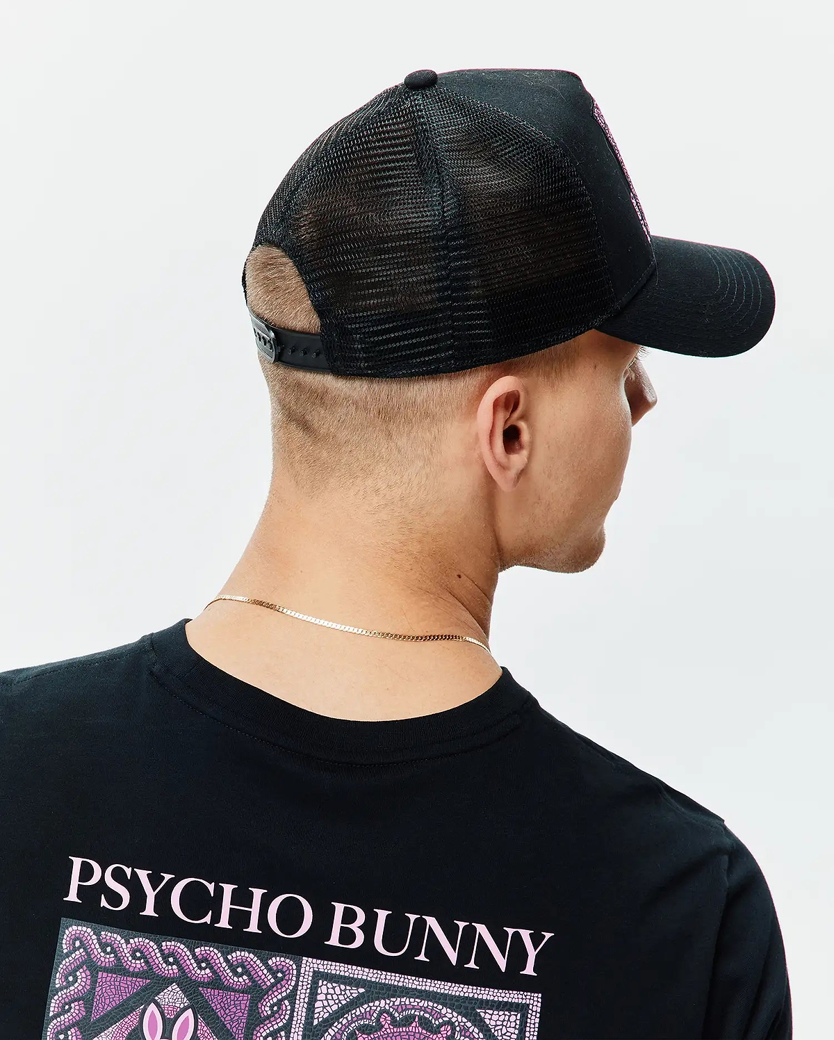 Black classic fit jersey t-shirt with large pink PSYCHO BUNNY text and mosaic graphic on the upper back, paired with a black mesh-back Mens Frank Trucker Cap featuring an embroidered pink Psycho Bunny logo on the front.
