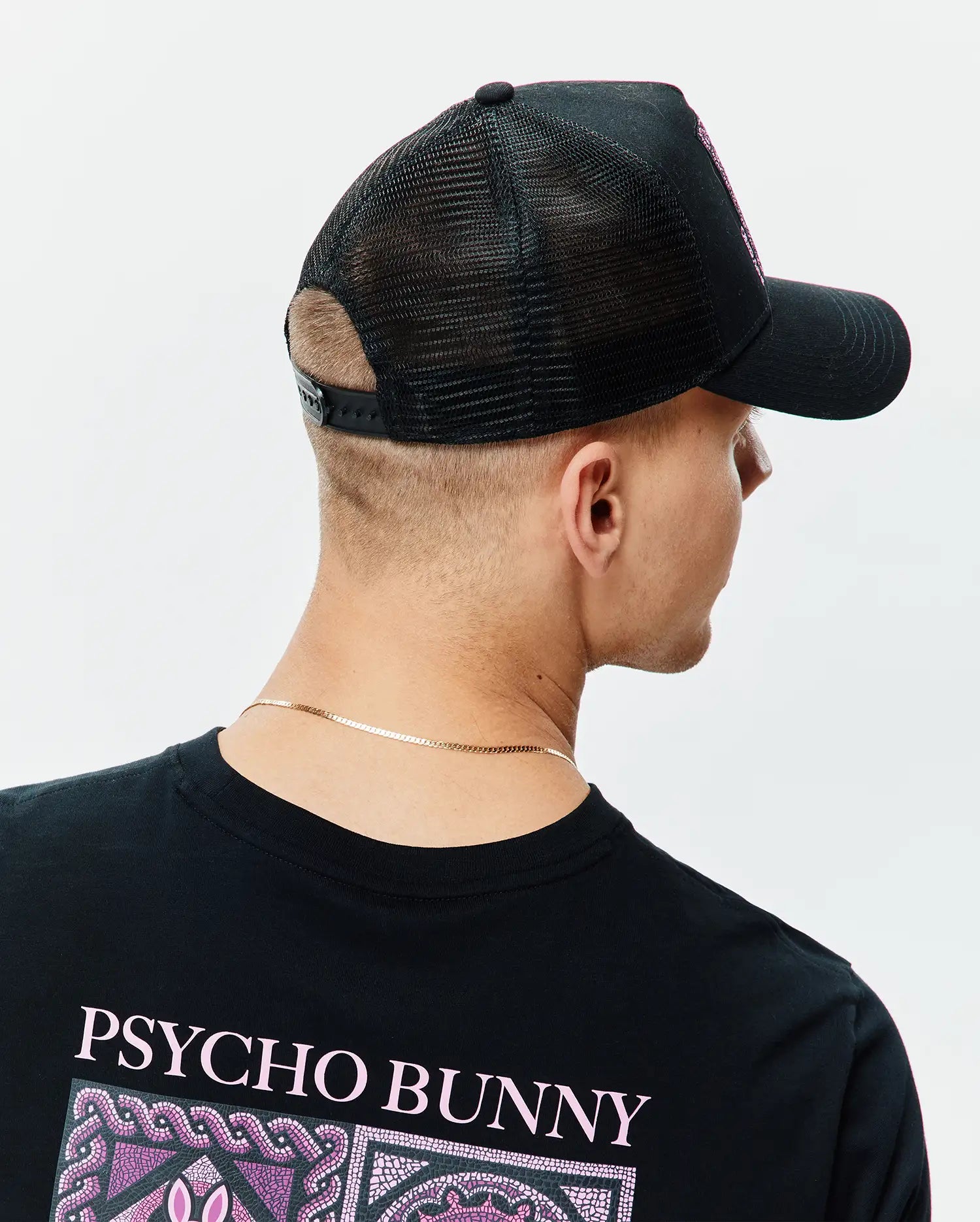 Black classic fit jersey t-shirt with large pink PSYCHO BUNNY text and mosaic graphic on the upper back, paired with a black mesh-back Mens Frank Trucker Cap featuring an embroidered pink Psycho Bunny logo on the front.