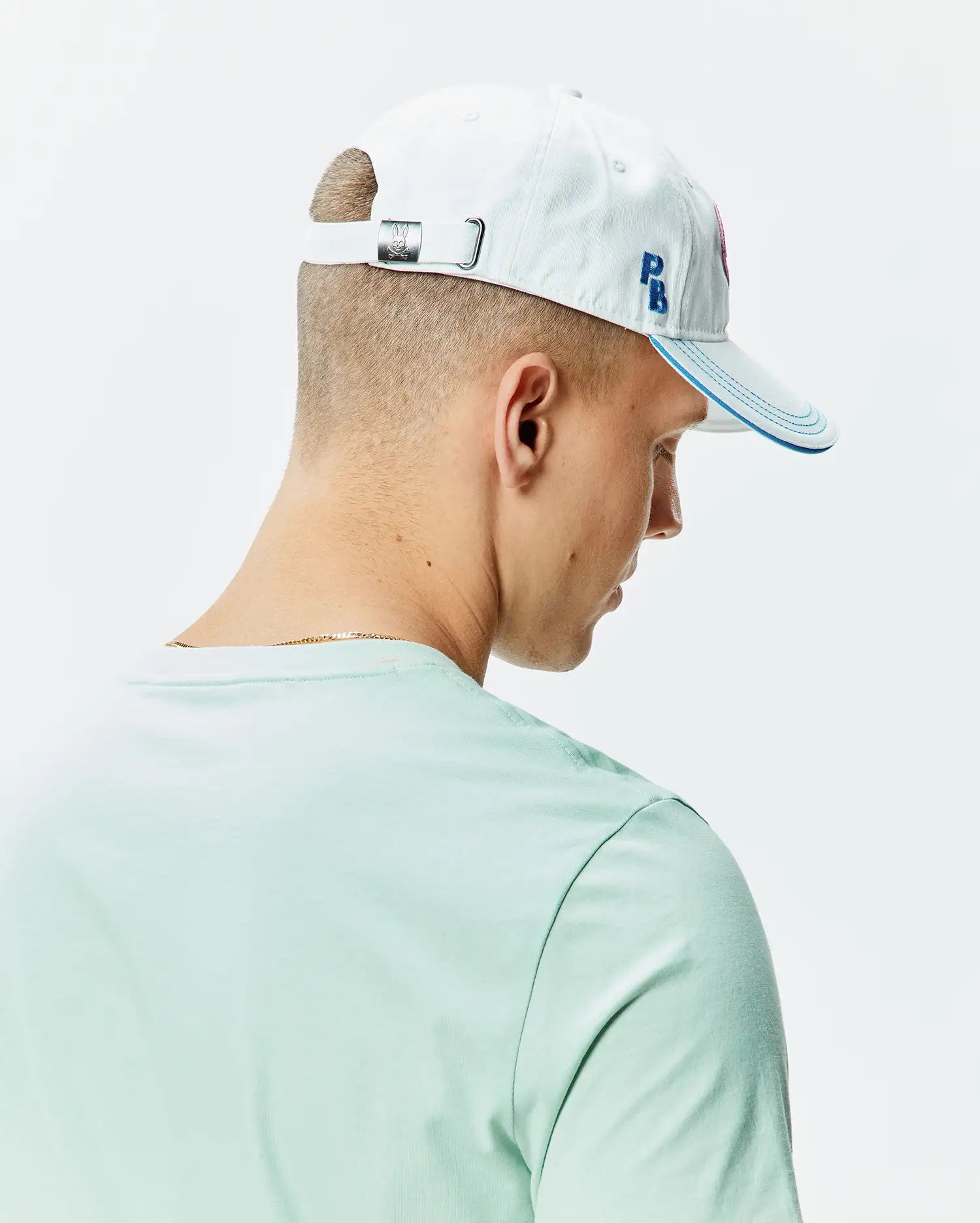 White cotton twill Mens Ray Baseball Cap with blue embroidered PB initials, blue brim stitching, and a metal Psycho Bunny skull buckle, paired with a mint green jersey knit crew neck t-shirt.