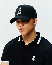 Black pique polo with white and grey striped collar and cuffs, featuring a small embroidered Psycho Bunny logo, paired with a black Mens Elijah Trucker Cap with a large white embroidered Psycho Bunny skull logo and mesh back.