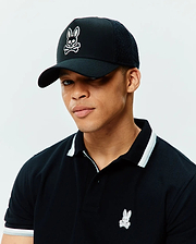 Black pique polo with white and grey striped collar and cuffs, featuring a small embroidered Psycho Bunny logo, paired with a black Mens Elijah Trucker Cap with a large white embroidered Psycho Bunny skull logo and mesh back.