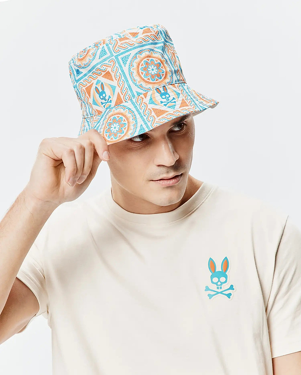 Mens Frank Reversible Bucket Hat in white with blue and orange mandala-style patterns featuring repeated Psycho Bunny skull logos, paired with a cream jersey t-shirt displaying a large blue and orange Psycho Bunny skull logo on the left chest.