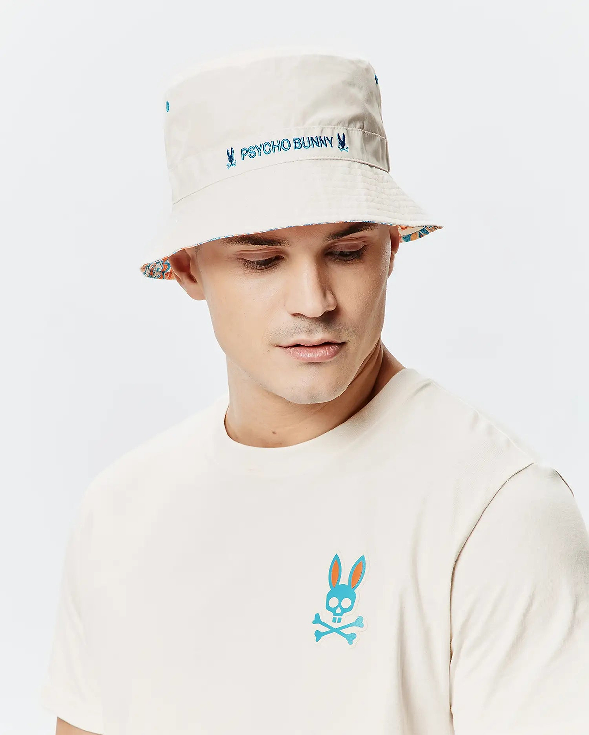 Mens Frank Reversible Bucket Hat in beige with blue Psycho Bunny embroidery and a colorful patterned reverse, paired with an off-white jersey t-shirt featuring a large blue and orange Psycho Bunny skull logo on the chest.