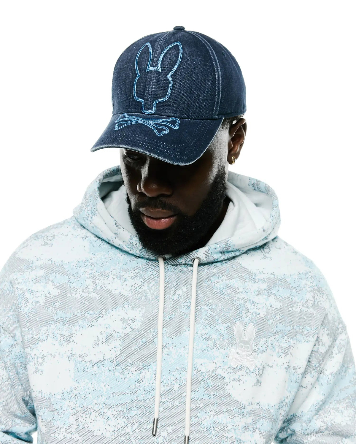Men's Grover Baseball Cap in dark denim with light blue embroidered Psycho Bunny logo, paired with a relaxed fit french terry hoodie featuring a light blue digital camo print and a small white embroidered Psycho Bunny chest logo. (1)