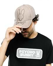 Men's Grover Baseball Cap in beige with dark brown embroidered Psycho Bunny skull and crossed bones, paired with a black classic fit jersey t-shirt featuring a large white textured Psycho Bunny logo on the chest. (1)