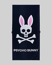 The Psycho Bunny MENS CLASSIC BUNNY TOWEL (MO1700004) is a dark towel with a white skull and crossbones topped by pink bunny ears above bold "PSYCHO BUNNY" lettering.