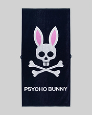The Psycho Bunny MENS CLASSIC BUNNY TOWEL (MO1700004) is a dark towel with a white skull and crossbones topped by pink bunny ears above bold "PSYCHO BUNNY" lettering.