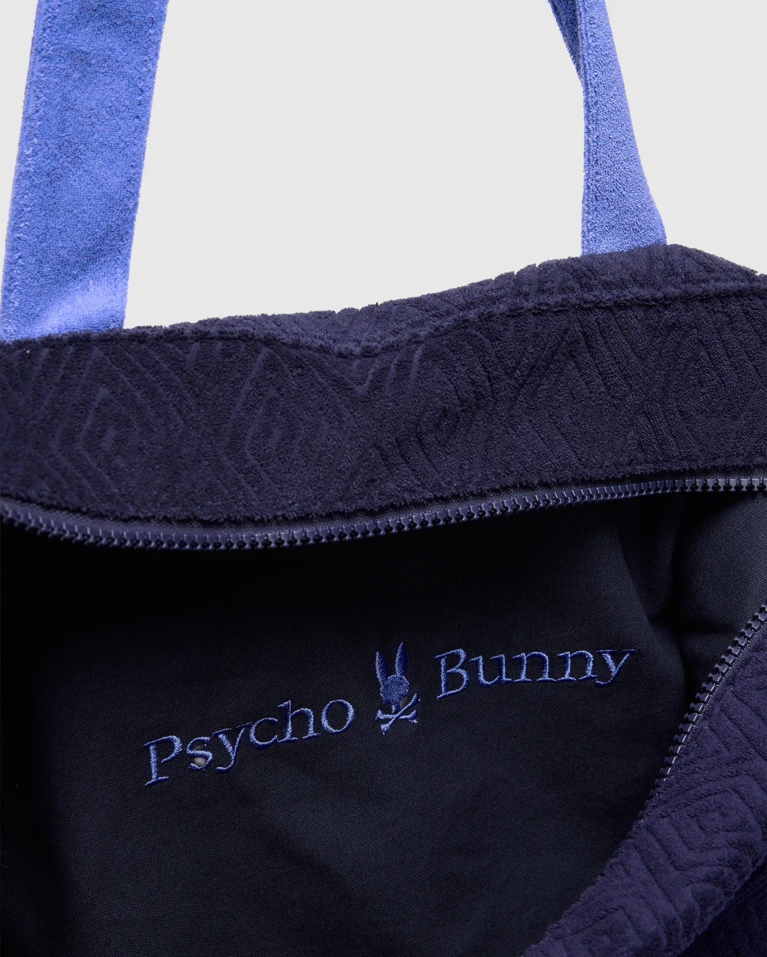 Navy Tristan Towel Tote with light blue terry straps, featuring an embroidered light blue Psycho Bunny skull logo and text on the smooth interior, and a subtle geometric pattern on the terry exterior.