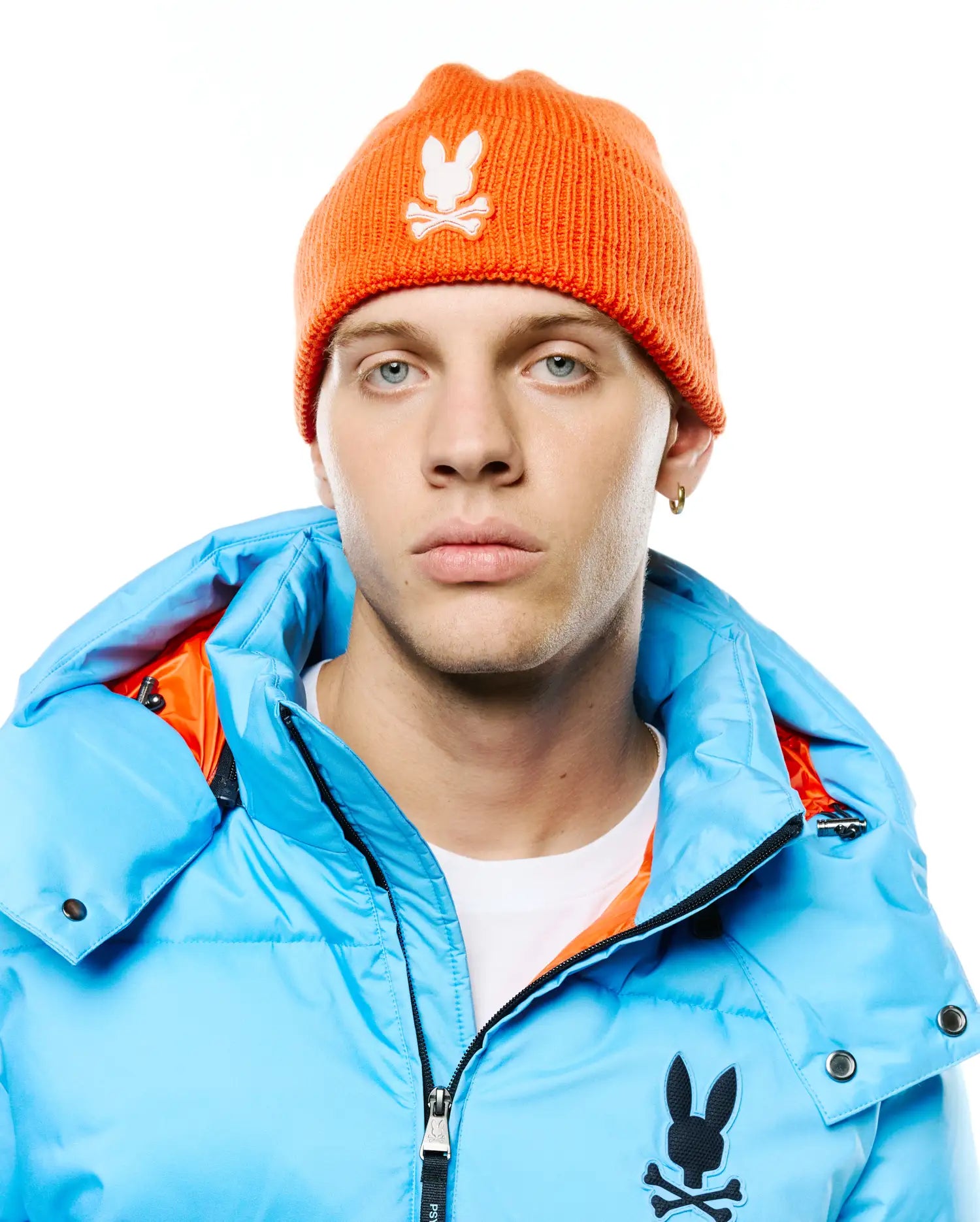 Bright orange ribbed knit Men's Leighton Merino Beanie with white embroidered Psycho Bunny logo, paired with a light blue puffer jacket featuring an orange lining and a large black embroidered Psycho Bunny logo on the chest. (1)