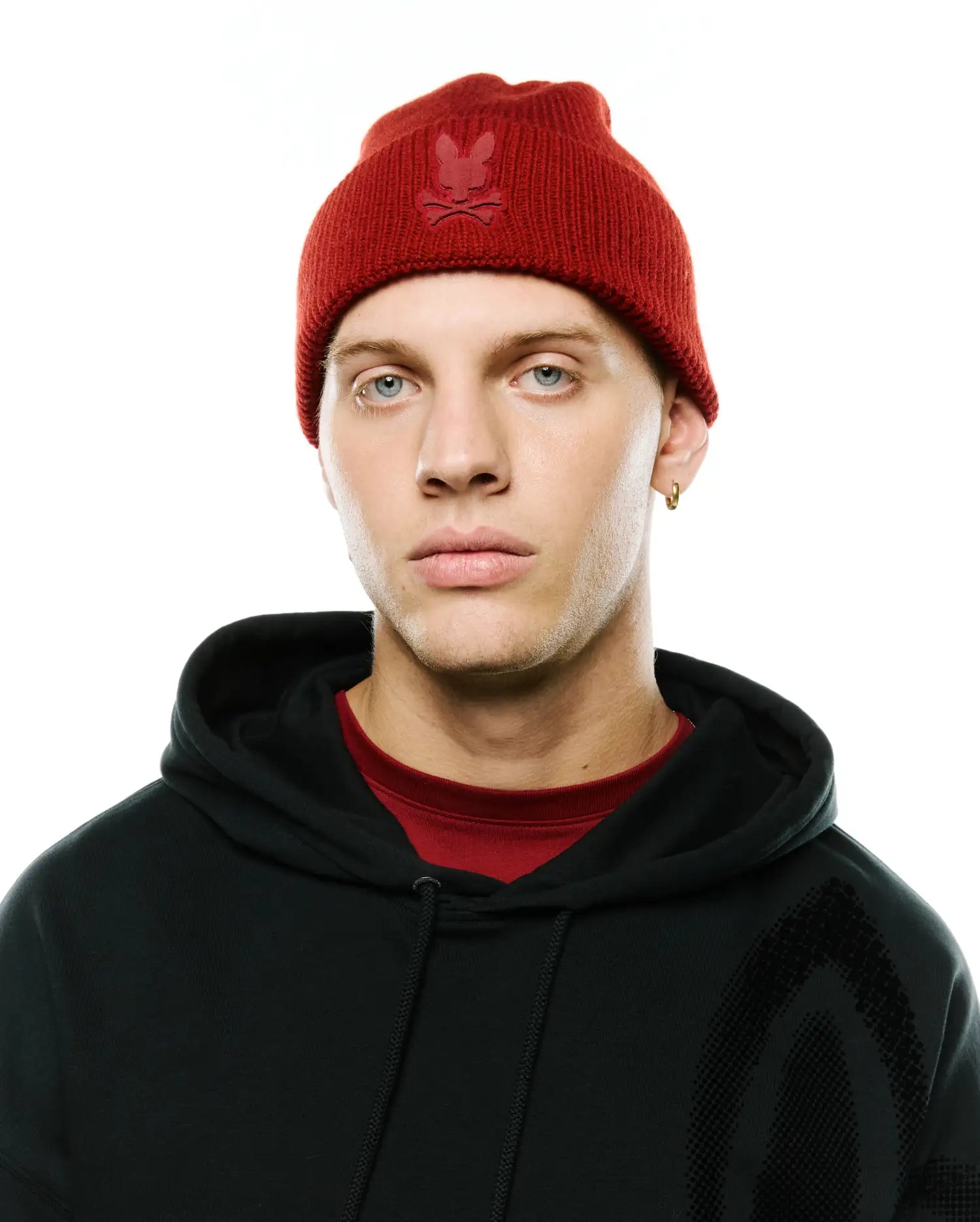 Red ribbed knit Men's Leighton Merino Beanie with tonal embroidered Psycho Bunny skull logo, paired with a black relaxed fit French terry hoodie featuring a subtle graphic print and a red crewneck t-shirt. (1)