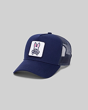 The Psycho Bunny MENS TRUCKER CAP - MO2600001 is a navy blue cap with a mesh back and a front patch showing a pink and white bunny above crossed bones, creating a playful skull and crossbones look, set against a plain gray background.