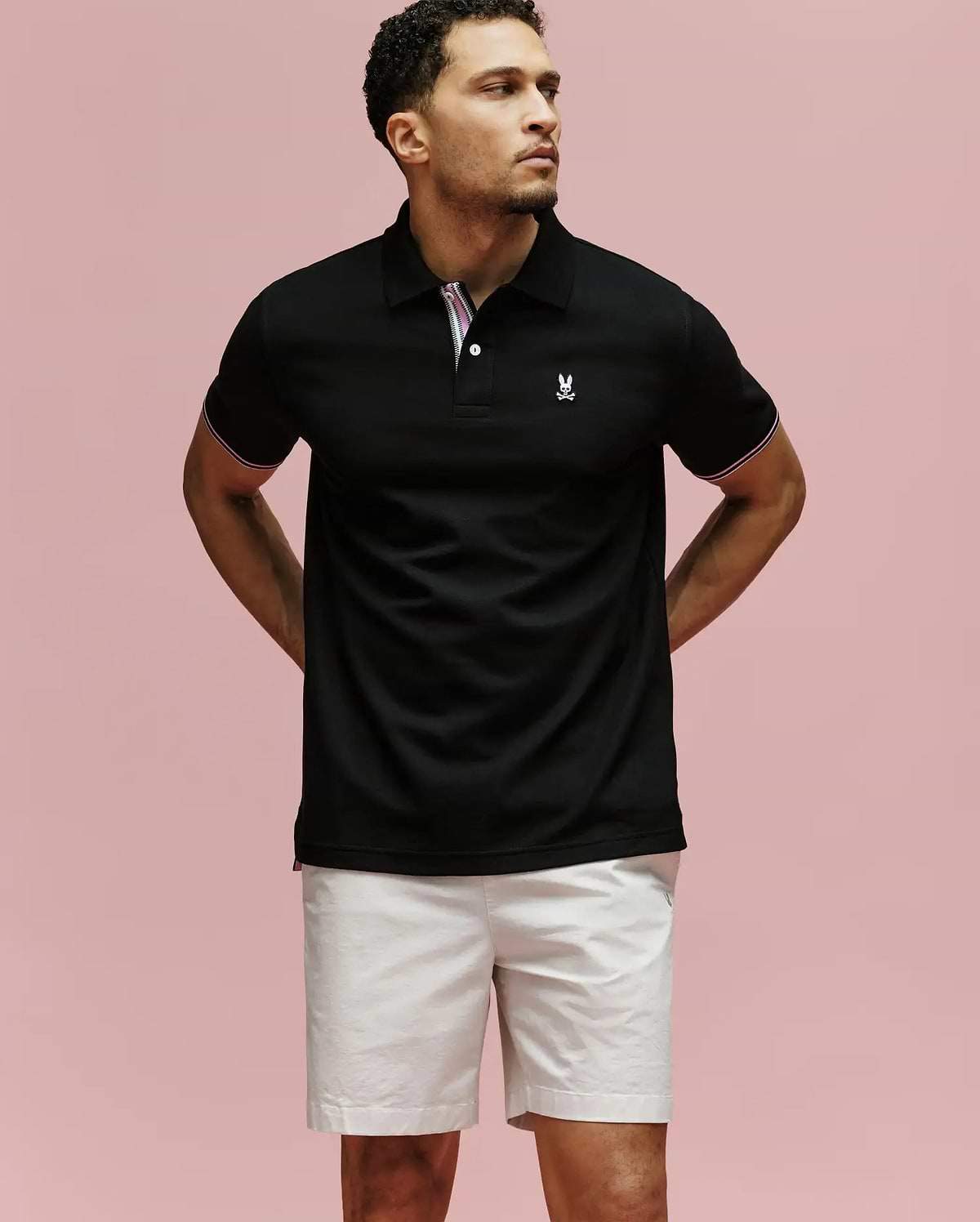 A man stands against a pink background, wearing the Psycho Bunny MENS ARCHIVE CLASSIC BUNNY PIQUE POLO (MT0100010) in black with an embroidered logo on the chest and light shorts. His arms are behind his back as he gazes to the side.