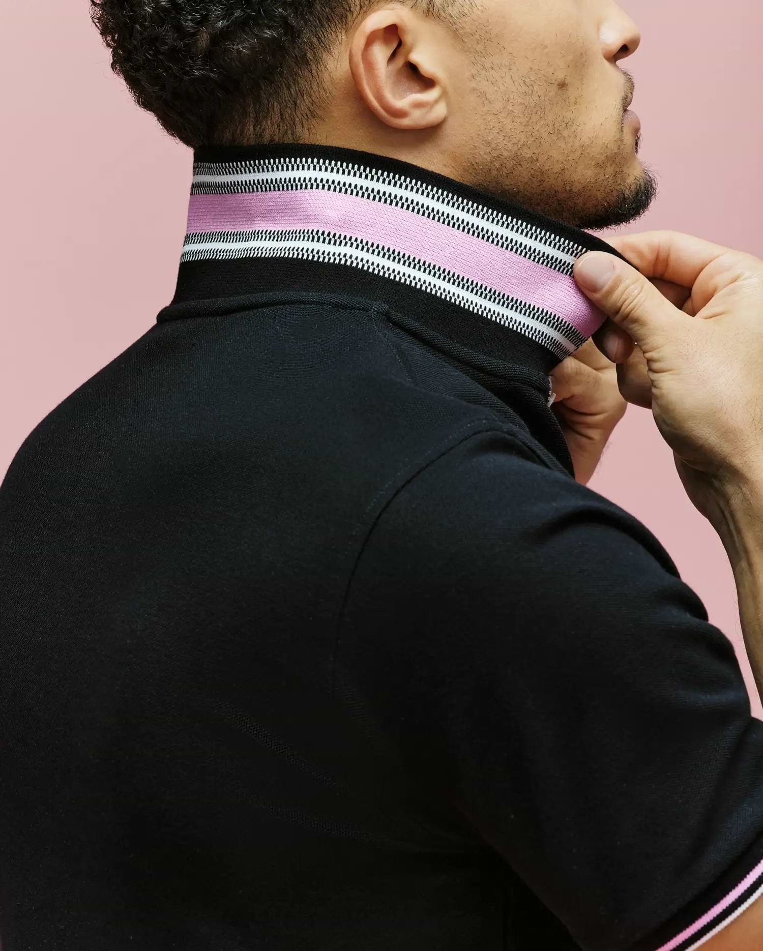 A person adjusts the high collar of Psycho Bunny's MENS ARCHIVE CLASSIC BUNNY PIQUE POLO (MT0100010), a black shirt with pink, white, and black stripes and an intarsia-knit undercollar, against a pink background. Only their lower face and upper torso are visible.