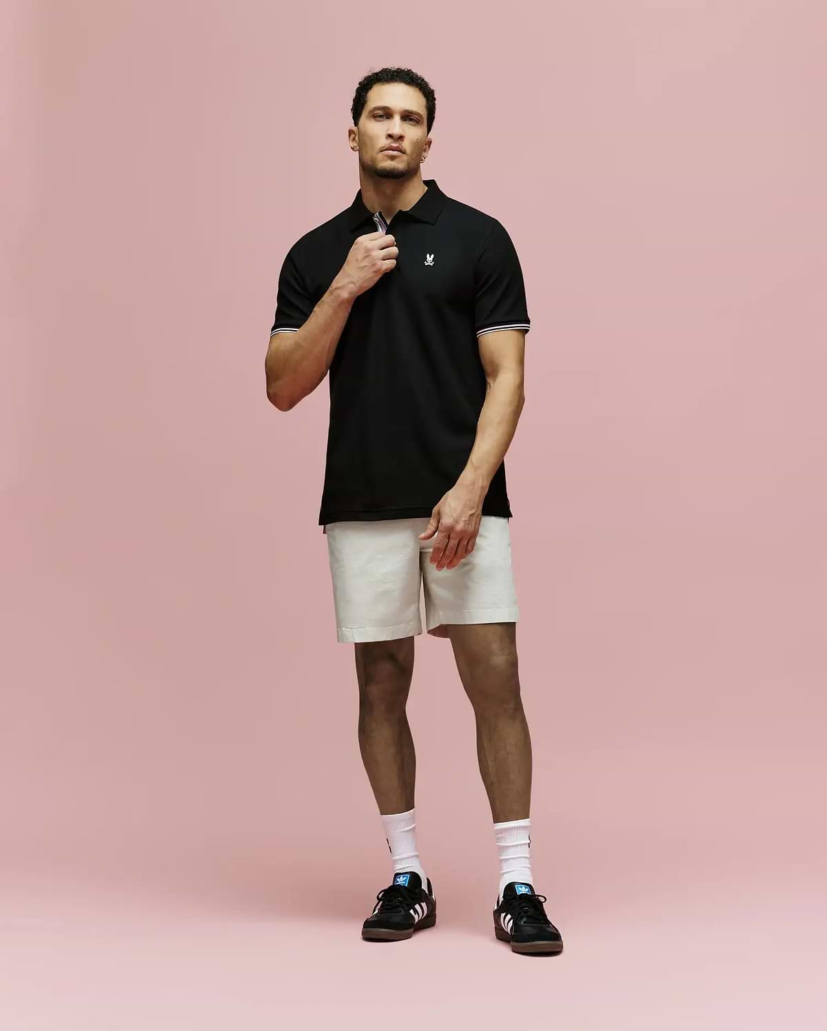 A man stands against a pink background wearing Psycho Bunny’s MENS ARCHIVE CLASSIC BUNNY PIQUE POLO - MT0100010, light beige shorts, white socks, and black Adidas sneakers. He holds the polo’s collar with one hand and looks forward.