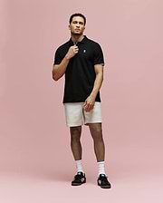 A man stands against a pink background wearing Psycho Bunny’s MENS ARCHIVE CLASSIC BUNNY PIQUE POLO - MT0100010, light beige shorts, white socks, and black Adidas sneakers. He holds the polo’s collar with one hand and looks forward.