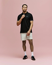 A man stands against a pink background wearing Psycho Bunny’s MENS ARCHIVE CLASSIC BUNNY PIQUE POLO - MT0100010, light beige shorts, white socks, and black Adidas sneakers. He holds the polo’s collar with one hand and looks forward.