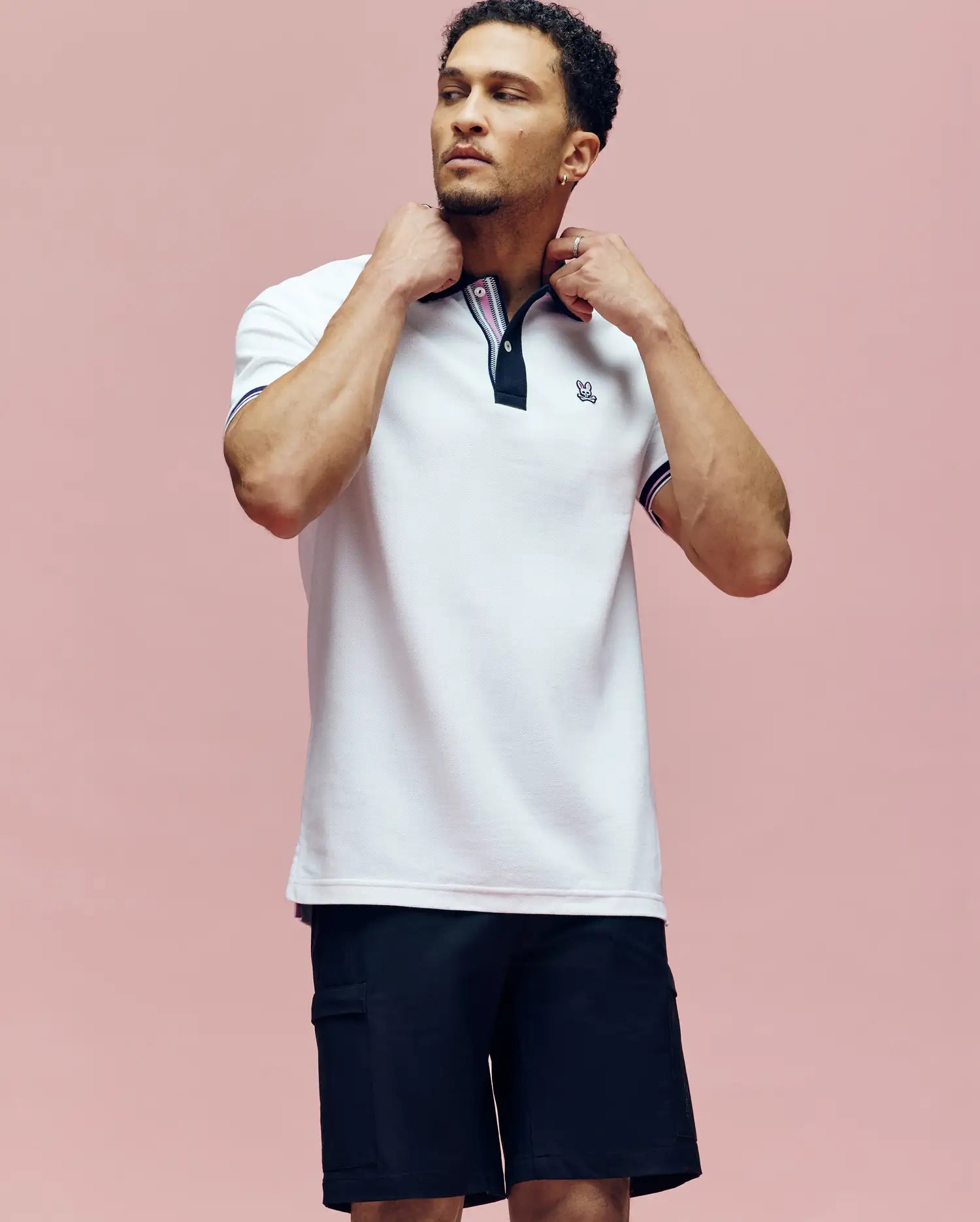 A man stands against a pink background, adjusting the collar of his white Psycho Bunny MENS ARCHIVE CLASSIC BUNNY PIQUE POLO (MT0100010) with dark trim. He is also wearing black shorts and gazing slightly upwards.