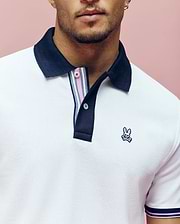 A man wears the Psycho Bunny MENS ARCHIVE CLASSIC BUNNY PIQUE POLO (MT0100010), a white Pima cotton shirt with a black collar, striped placket, and embroidered Bunny skull logo, standing against a pink background.