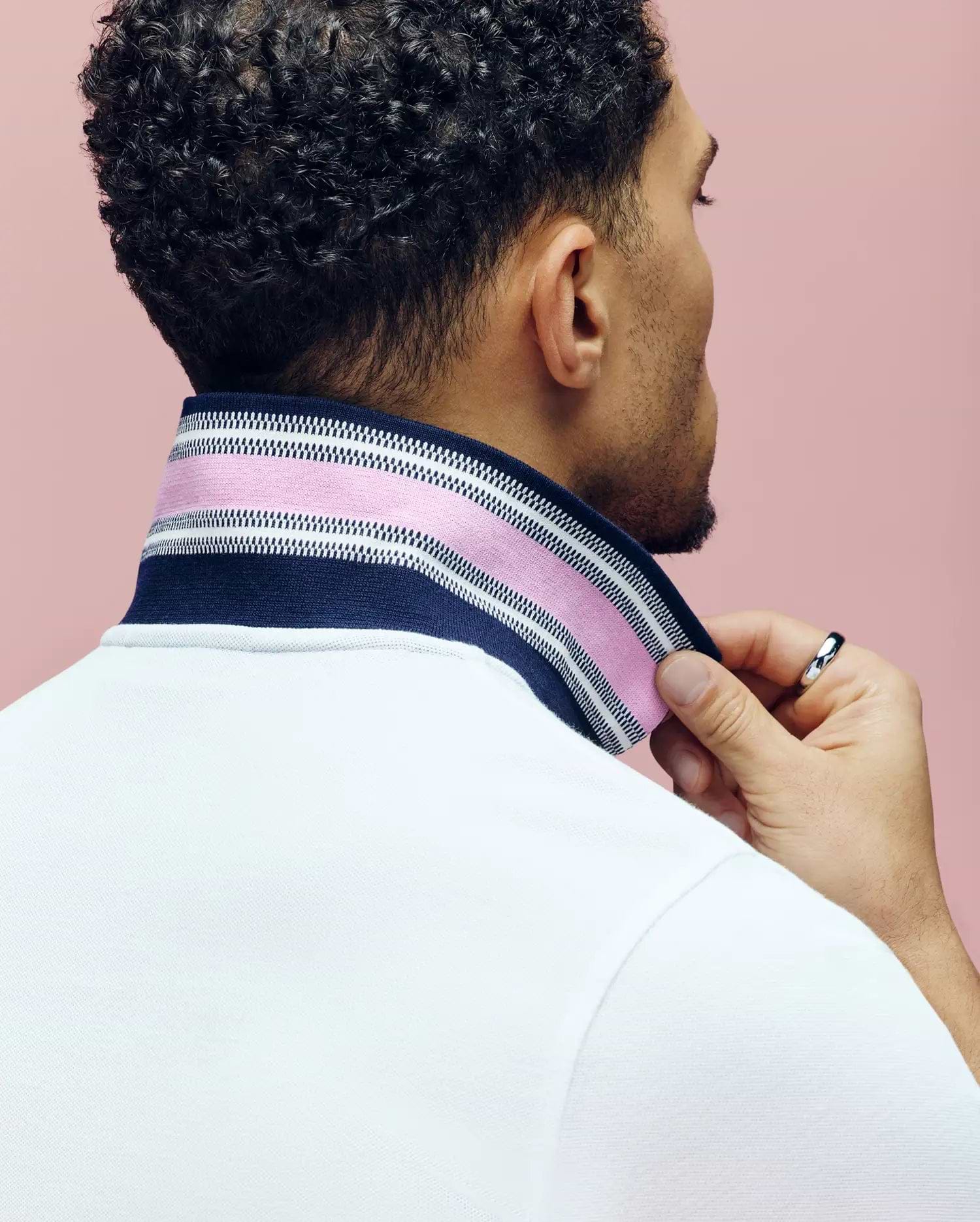 A man with curly hair, seen from behind, lifts the collar of his white Psycho Bunny MENS ARCHIVE CLASSIC BUNNY PIQUE POLO to reveal a striped navy, pink, and white pattern. He wears a silver ring and stands against a pink background.