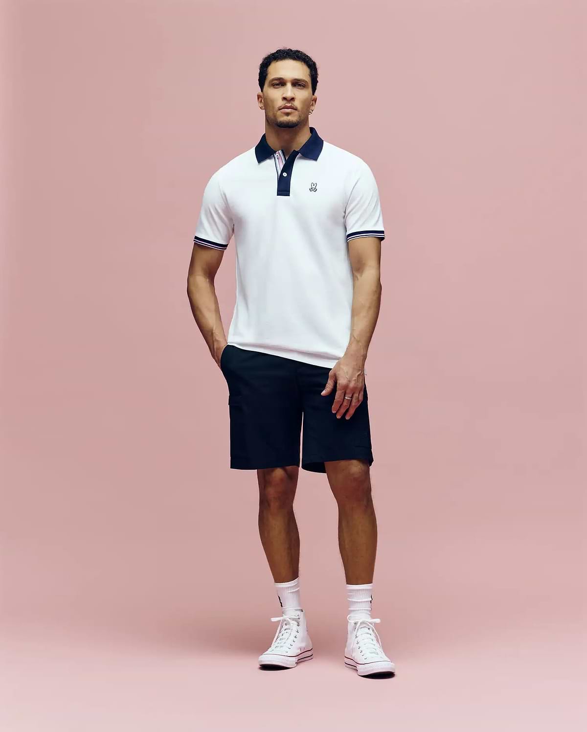 A man stands against a pink background wearing the Psycho Bunny MENS ARCHIVE CLASSIC BUNNY PIQUE POLO - MT0100010, featuring white Pima cotton with a dark collar, paired with black shorts, white socks, and high-top sneakers.
