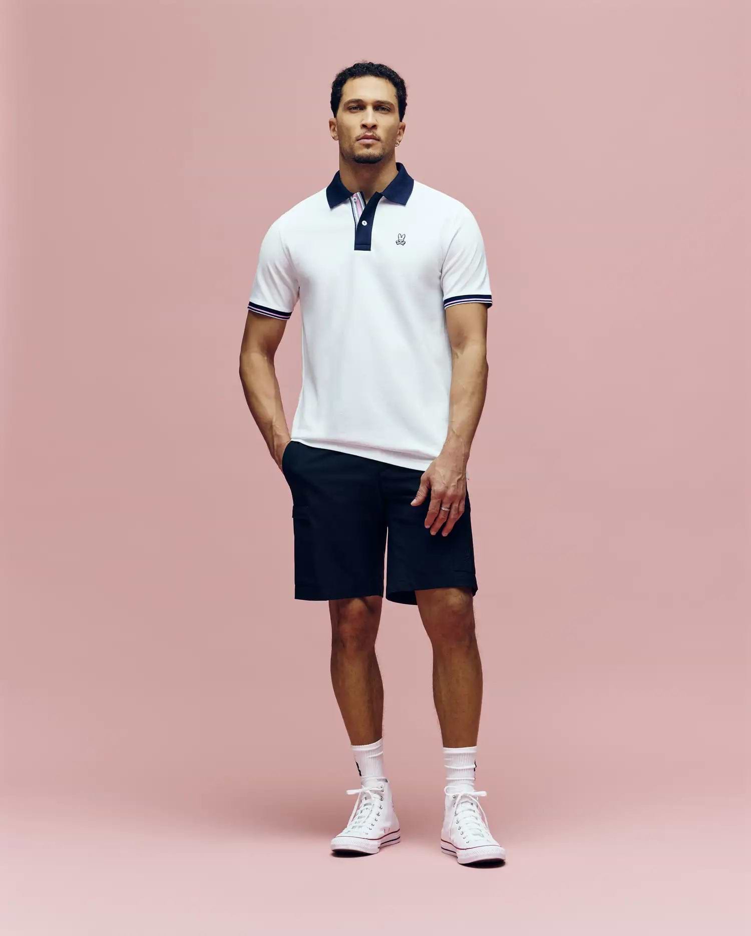 A man stands against a pink background wearing the Psycho Bunny MENS ARCHIVE CLASSIC BUNNY PIQUE POLO - MT0100010, featuring white Pima cotton with a dark collar, paired with black shorts, white socks, and high-top sneakers.