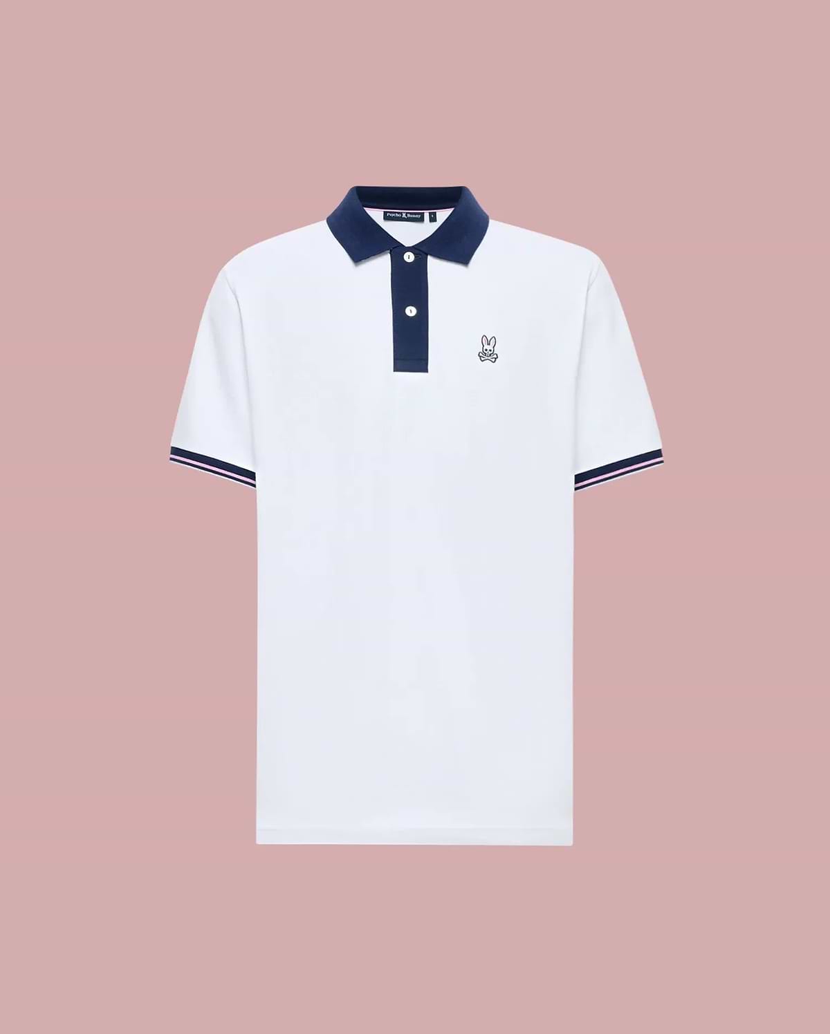 The Psycho Bunny MENS ARCHIVE CLASSIC BUNNY PIQUE POLO (MT0100010) is a white Pima cotton short-sleeve polo with a navy collar, button placket, sleeve trim, and an embroidered Bunny logo on the chest, shown on a pink background.