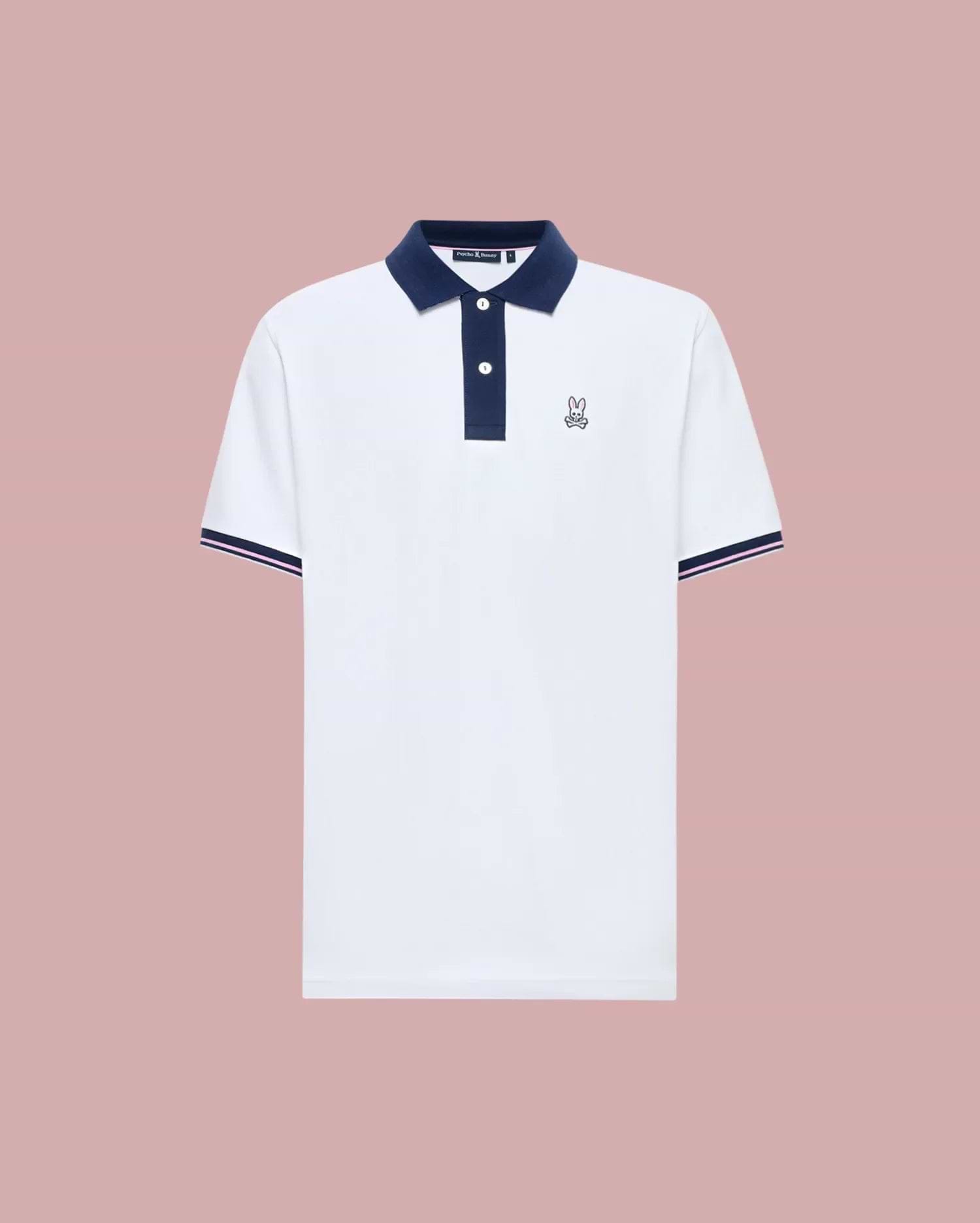 The Psycho Bunny MENS ARCHIVE CLASSIC BUNNY PIQUE POLO (MT0100010) is a white Pima cotton short-sleeve polo with a navy collar, button placket, sleeve trim, and an embroidered Bunny logo on the chest, shown on a pink background.