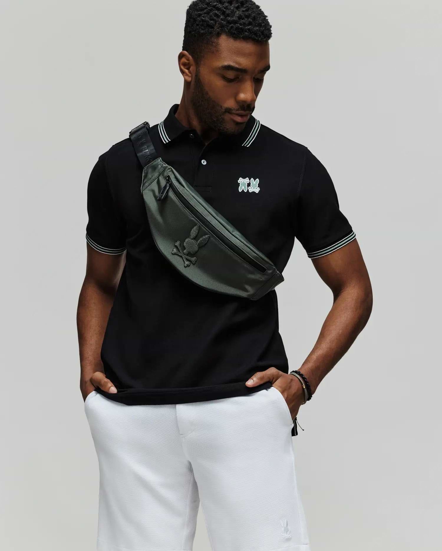 A man stands against a plain background wearing the Psycho Bunny MENS ARCHIVE NEON BUNNY POLO (MT0100018) in black with white trim, paired with white shorts and a green crossbody bag. The shirt features an embroidered Bunny detail. He looks down, hands in pockets. (1)