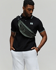 A man stands against a plain background wearing the Psycho Bunny MENS ARCHIVE NEON BUNNY POLO (MT0100018) in black with white trim, paired with white shorts and a green crossbody bag. The shirt features an embroidered Bunny detail. He looks down, hands in pockets. (1)