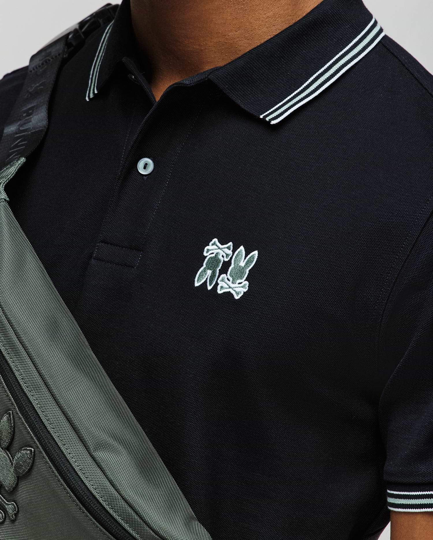 A person wearing a Psycho Bunny MENS ARCHIVE NEON BUNNY POLO (MT0100018) in black with white trim and an embroidered Bunny logo, paired with a grey crossbody bag featuring the same logo. Only the upper torso is visible. (2)