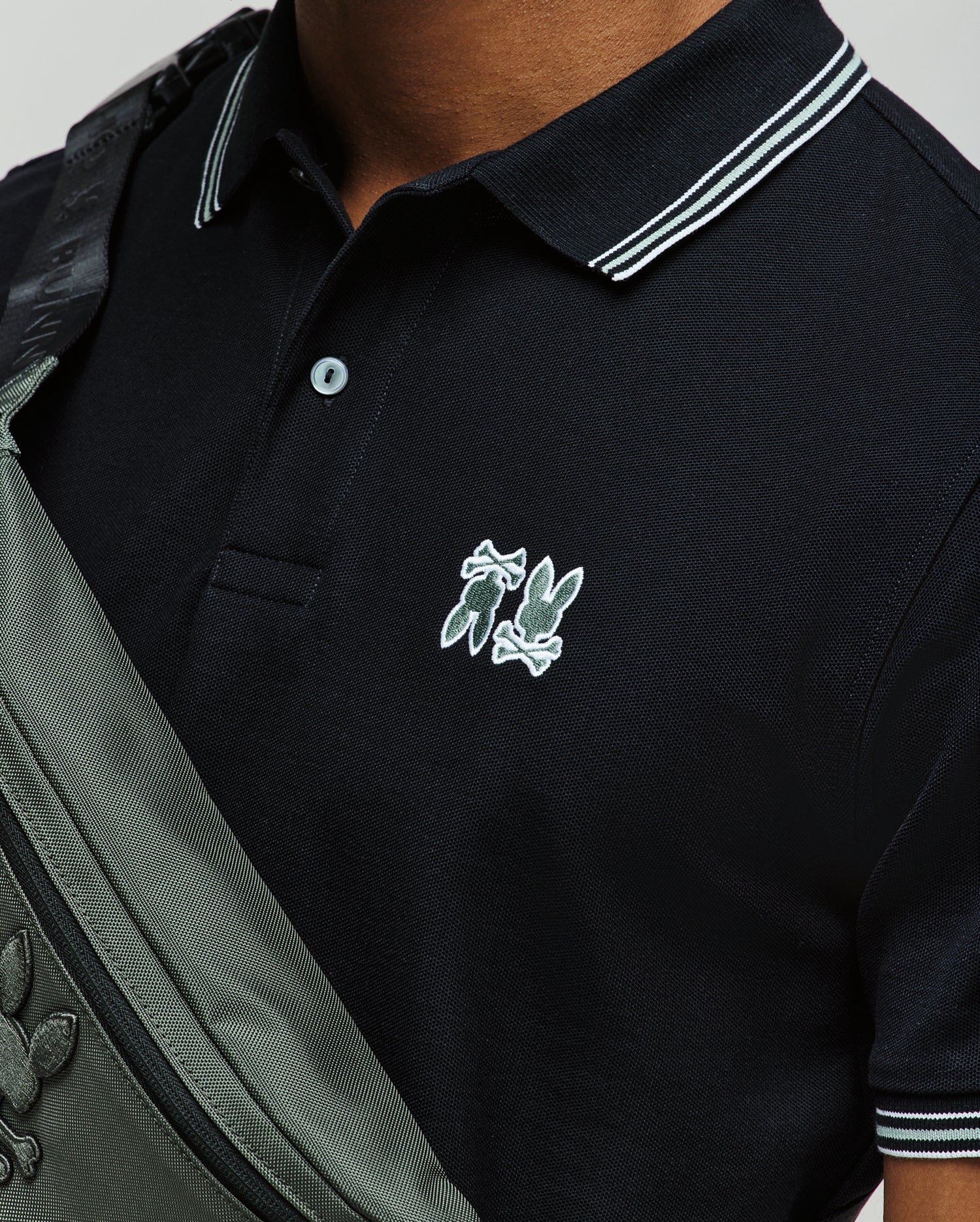 A person wearing a Psycho Bunny MENS ARCHIVE NEON BUNNY POLO (MT0100018) in black with white trim and an embroidered Bunny logo, paired with a grey crossbody bag featuring the same logo. Only the upper torso is visible. (2)
