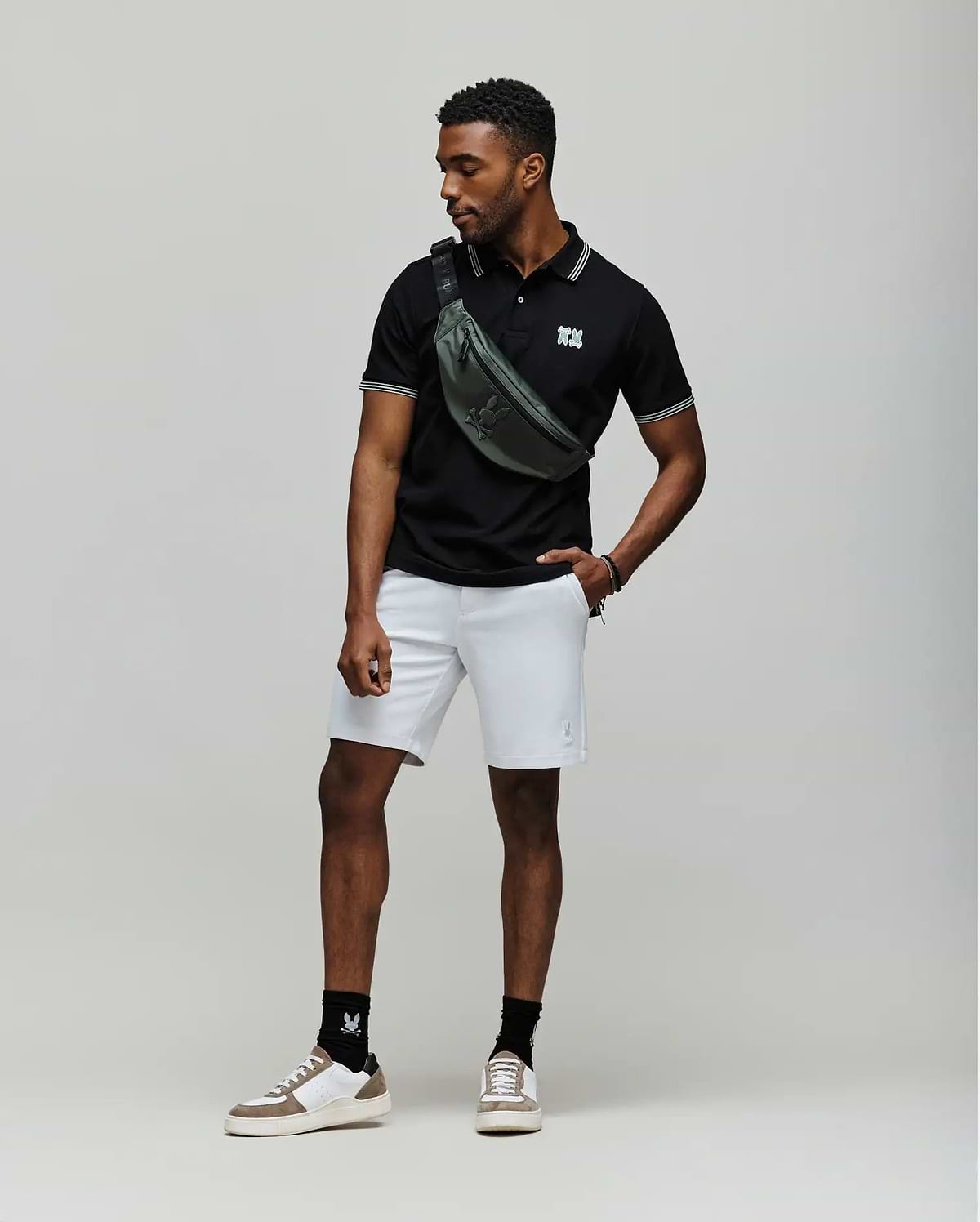 A man stands against a plain backdrop wearing a black Psycho Bunny MENS ARCHIVE NEON BUNNY POLO - MT0100018 made from 100% Pima cotton, light gray shorts, white sneakers with black socks, and a green crossbody bag. He looks to the side, one hand in his pocket. (3)