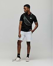 A man stands against a plain backdrop wearing a black Psycho Bunny MENS ARCHIVE NEON BUNNY POLO - MT0100018 made from 100% Pima cotton, light gray shorts, white sneakers with black socks, and a green crossbody bag. He looks to the side, one hand in his pocket. (3)