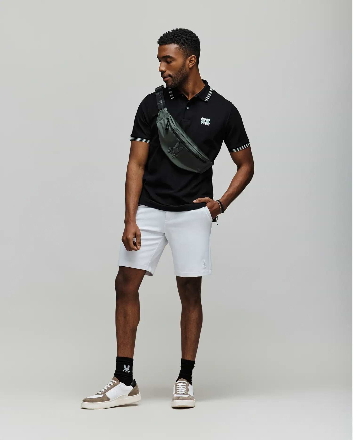 A man stands against a plain backdrop wearing a black Psycho Bunny MENS ARCHIVE NEON BUNNY POLO - MT0100018 made from 100% Pima cotton, light gray shorts, white sneakers with black socks, and a green crossbody bag. He looks to the side, one hand in his pocket. (3)