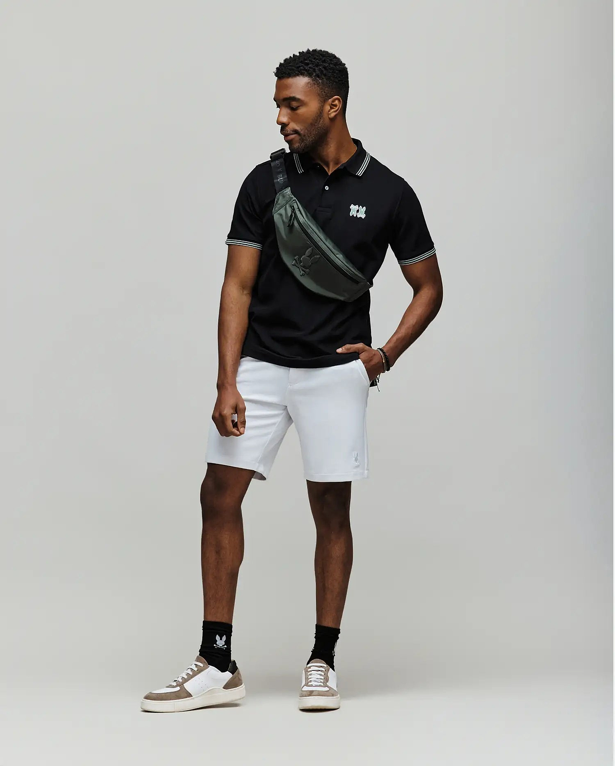 A man stands against a plain backdrop wearing a black Psycho Bunny MENS ARCHIVE NEON BUNNY POLO - MT0100018 made from 100% Pima cotton, light gray shorts, white sneakers with black socks, and a green crossbody bag. He looks to the side, one hand in his pocket. (3)