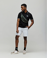 A man stands against a plain backdrop wearing a black Psycho Bunny MENS ARCHIVE NEON BUNNY POLO - MT0100018 made from 100% Pima cotton, light gray shorts, white sneakers with black socks, and a green crossbody bag. He looks to the side, one hand in his pocket. (3)