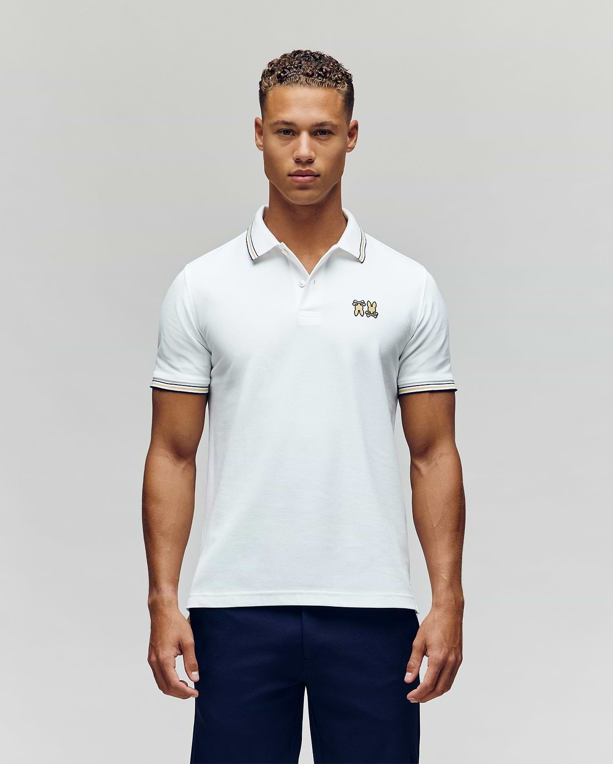 A man faces forward in dark pants and a Psycho Bunny MENS ARCHIVE NEON BUNNY POLO MT0100018—a regular fit white, 100% Pima cotton short-sleeve shirt featuring a small embroidered Bunny on the chest—against a plain light background. (4)