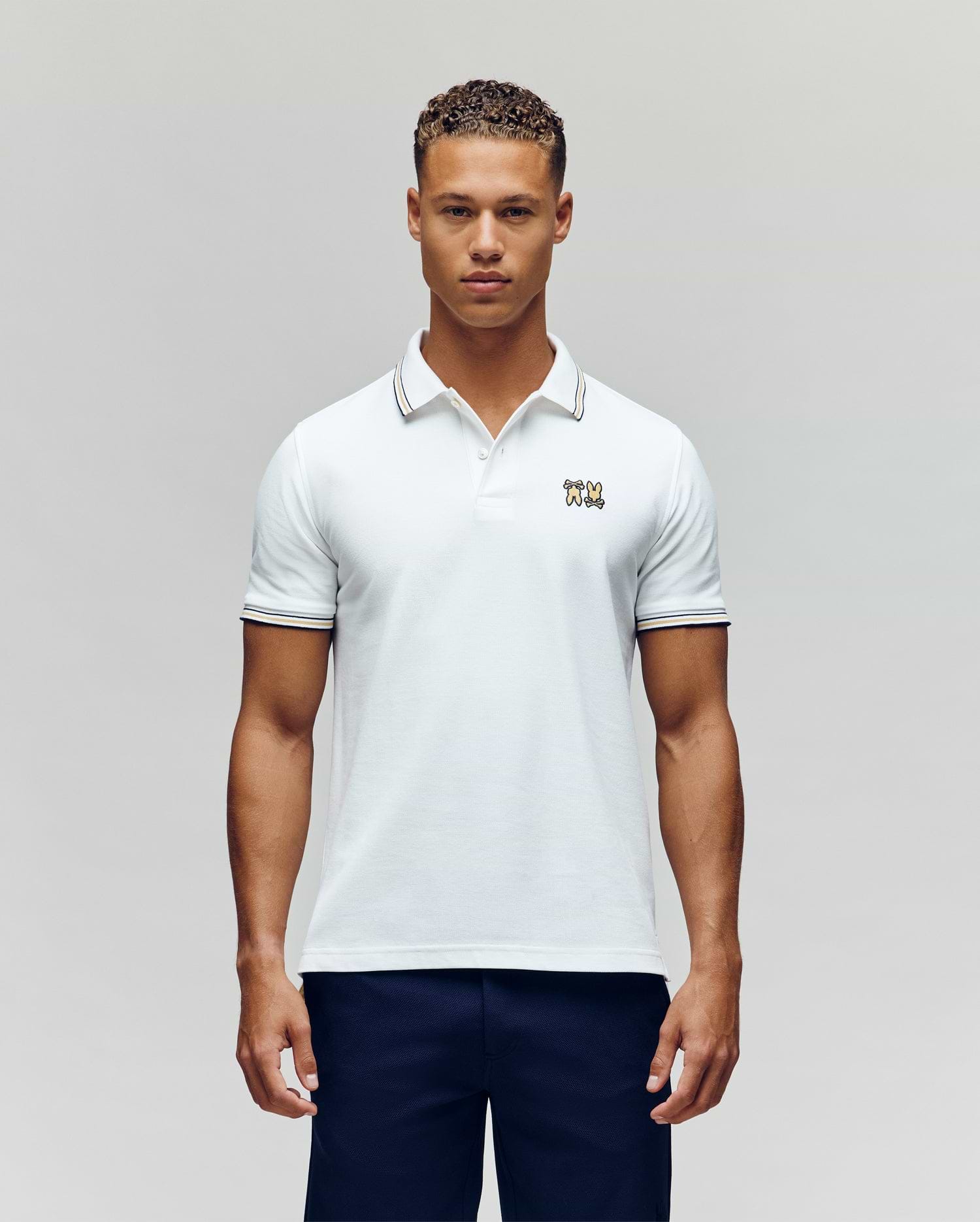 A man faces forward in dark pants and a Psycho Bunny MENS ARCHIVE NEON BUNNY POLO MT0100018—a regular fit white, 100% Pima cotton short-sleeve shirt featuring a small embroidered Bunny on the chest—against a plain light background. (4)