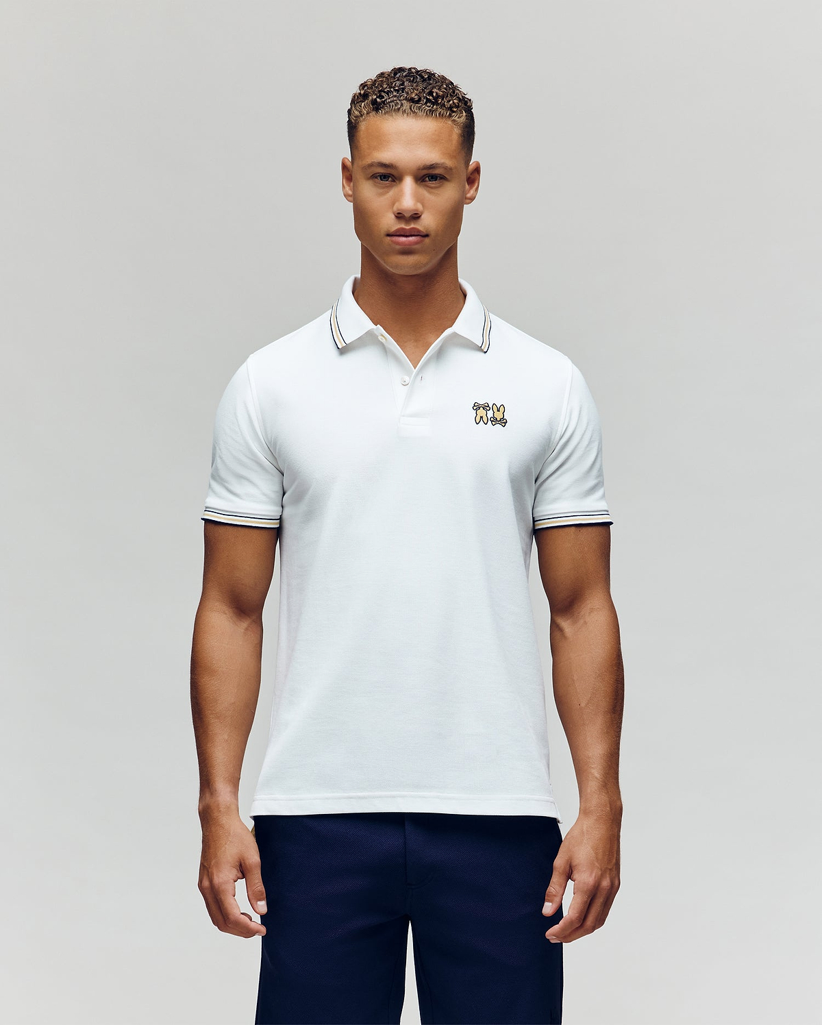 A man faces forward in dark pants and a Psycho Bunny MENS ARCHIVE NEON BUNNY POLO MT0100018—a regular fit white, 100% Pima cotton short-sleeve shirt featuring a small embroidered Bunny on the chest—against a plain light background. (4)
