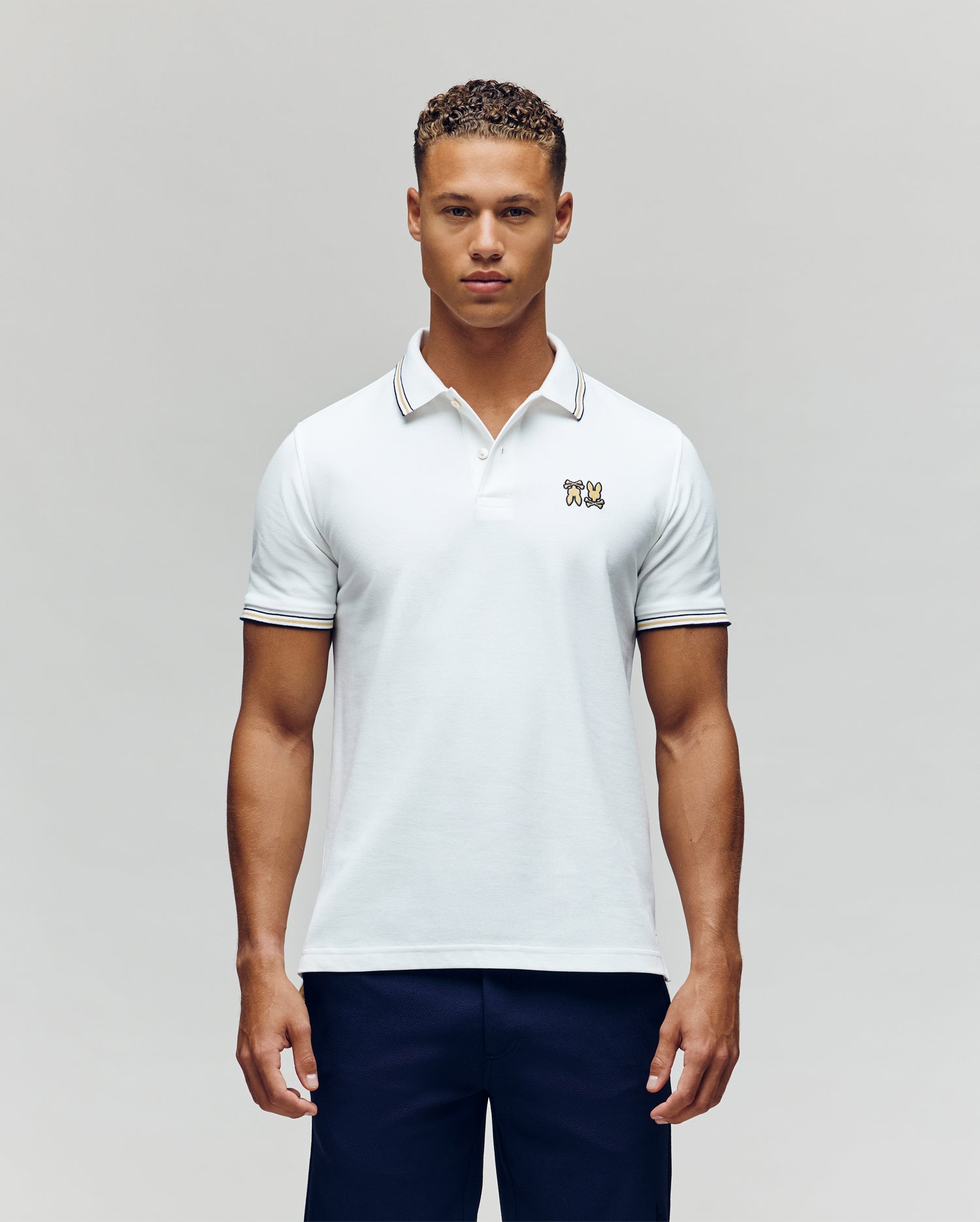 A man faces forward in dark pants and a Psycho Bunny MENS ARCHIVE NEON BUNNY POLO MT0100018—a regular fit white, 100% Pima cotton short-sleeve shirt featuring a small embroidered Bunny on the chest—against a plain light background. (4)