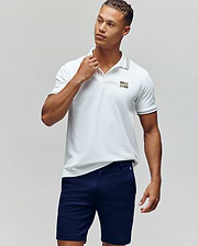 A man stands against a plain background in the Psycho Bunny MENS ARCHIVE NEON BUNNY POLO - MT0100018, a white, regular fit 100% Pima cotton shirt with an embroidered Bunny logo, paired with navy blue shorts. He looks to the side and touches his collar. (1)