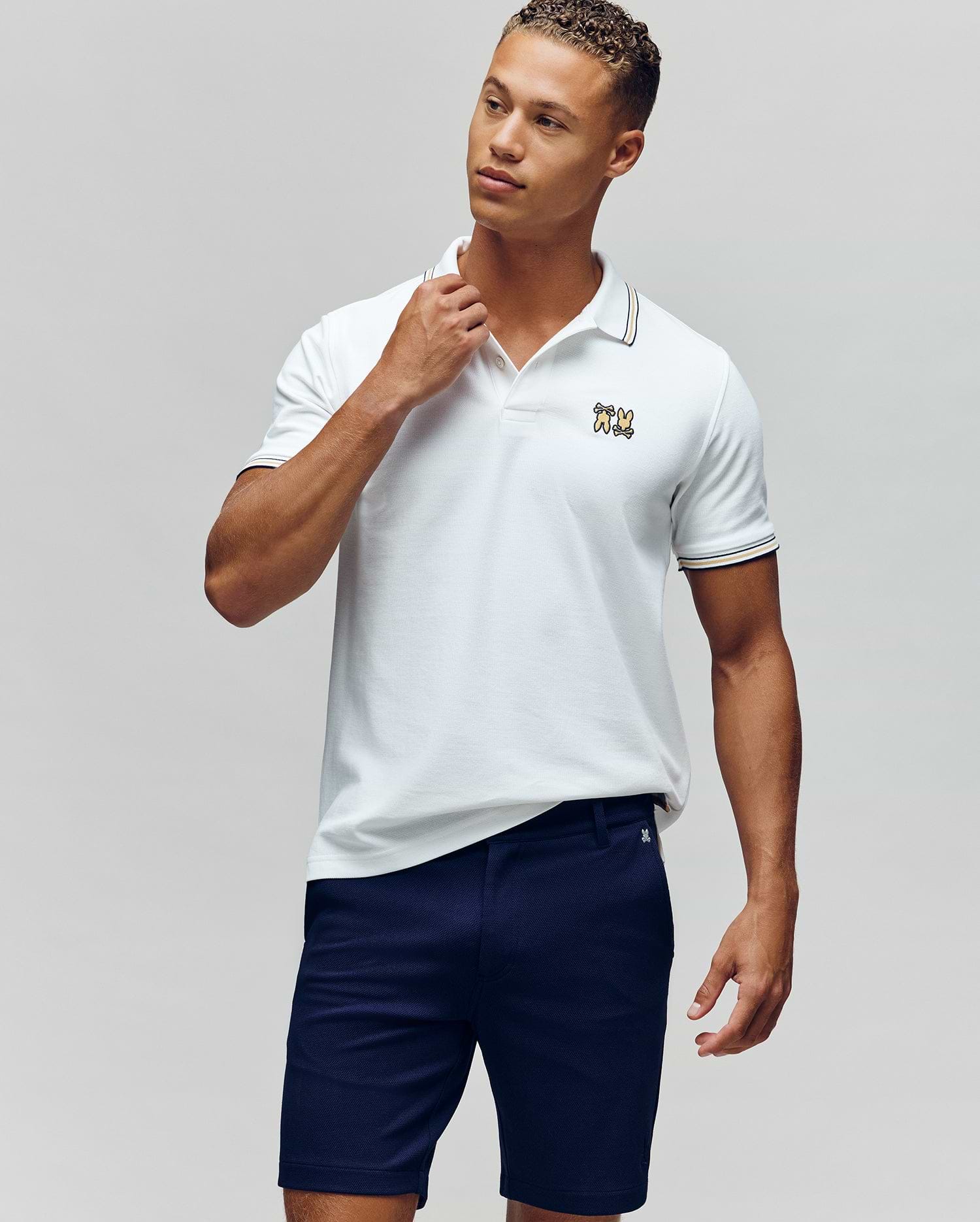 A man stands against a plain background in the Psycho Bunny MENS ARCHIVE NEON BUNNY POLO - MT0100018, a white, regular fit 100% Pima cotton shirt with an embroidered Bunny logo, paired with navy blue shorts. He looks to the side and touches his collar. (1)