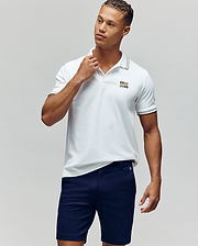 A man stands against a plain background in the Psycho Bunny MENS ARCHIVE NEON BUNNY POLO - MT0100018, a white, regular fit 100% Pima cotton shirt with an embroidered Bunny logo, paired with navy blue shorts. He looks to the side and touches his collar. (1)